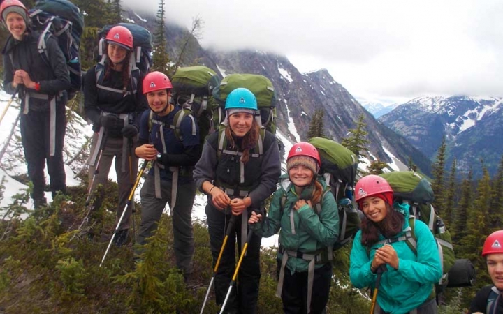 A group of people wearing backpacks and helmets smile at the camera. Behind them are snow covered mountains.